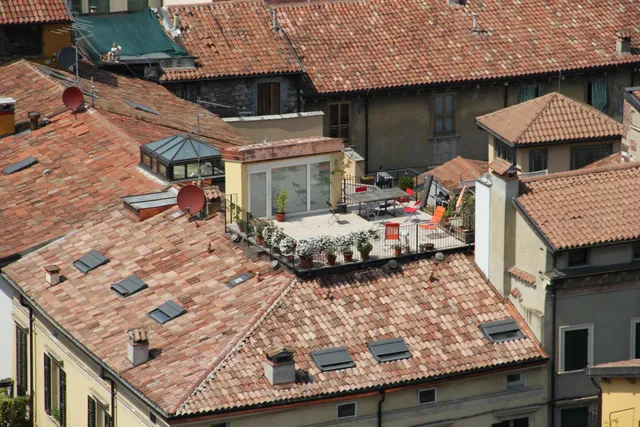 Tejados de tejas rojizas con chimeneas, ventanas de cubierta y antenas parabólicas alrededor de una terraza con macetas, mesas y sillas.